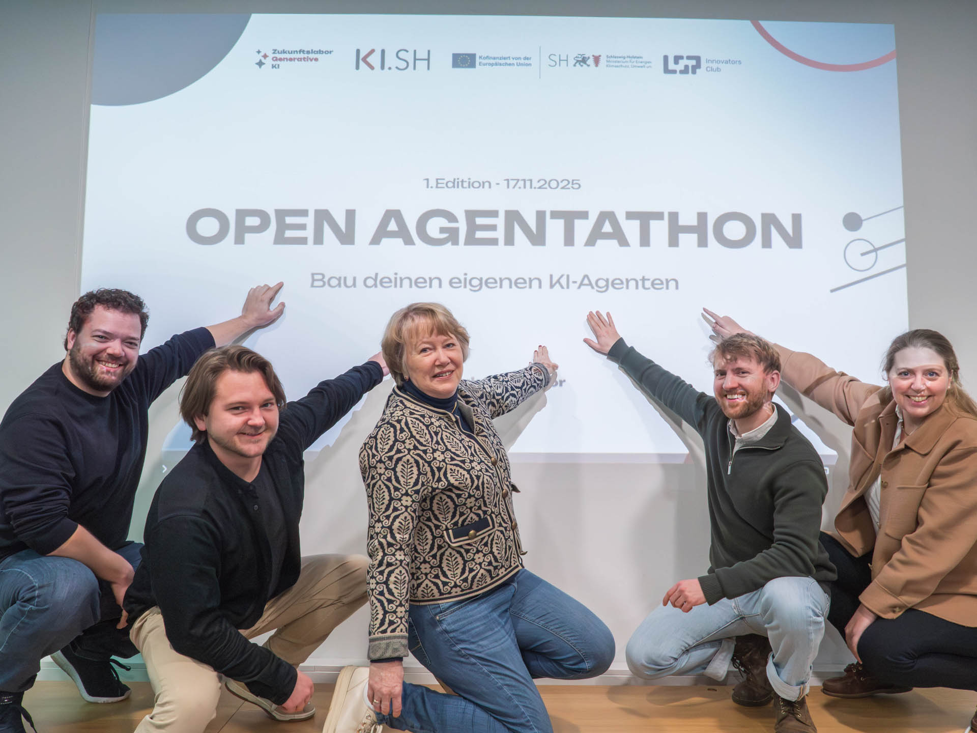 fünf Personen hocken vor einer Beamerleinwand und zeigen auf den Titel der Veranstaltung